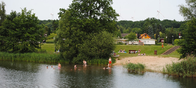 Die Menschen schwimmen und entspannen sich an einem kleinen Strandbereich an einem See, der von Gras, Bäumen und nahe gelegenen Häusern umgeben ist.